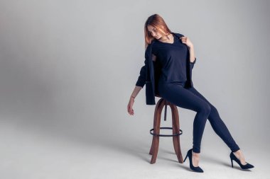 Koyu renk takım elbiseli ve yüksek topuklu ayakkabılı bir kız profilden bir sandalyede oturuyor ve başı eğik ve sırtüstü yatıyor.