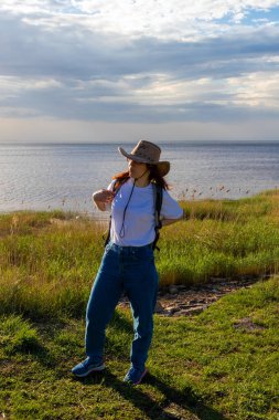 a girl in a hat puts a backpack on her back against the background of nature