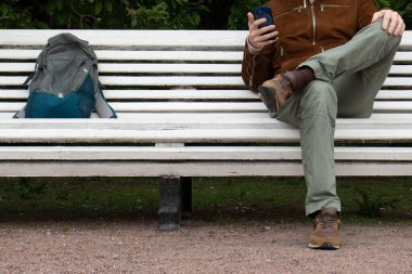 a man in a brown jacket and brown shoes is sitting on a white bench in the park in the afternoon with a phone in his hands. The backpack is lying next to him on a bench