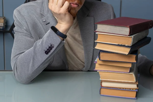 the man in the jacket has his head propped up and he\'s looking at a large stack of books. hand and books in the frame