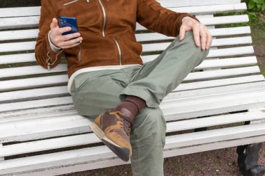 Şık giyimli genç bir adam parktaki beyaz bir bankta oturuyor ve elinde bir telefon var.