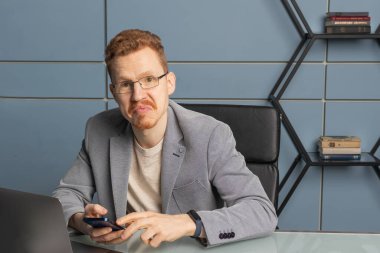 Kırmızı saçlı, gözlüklü ve gri ceketli bir işadamı masasında oturuyor.