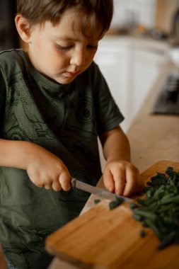 Evde, mutfakta beş yaşında bir çocuk yeşil soğanları bıçakla kesiyor. Çocuk yemek yapıyor.