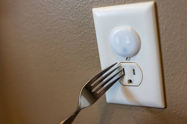 Selective focus on edge of a metal fork about to be inserted into an open electrical outlet socket. The top outlet has a plastic insert for childproofing safety protection.