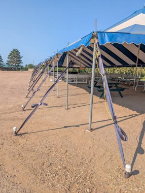 A large, blue and white striped tent is firmly secured with straps at a community event location. The area is clear, showing gravel ground and a few nearby benches under a sunny sky.