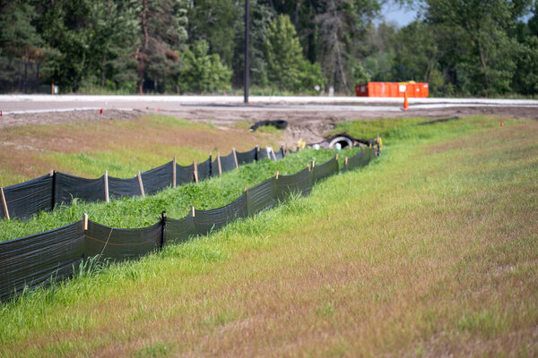 A construction area features a long black silt fence along the edge of a grassy field. The sun shines down, illuminating the surrounding greenery and trees in the distance.