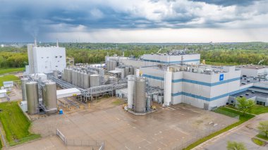 Büyük bir sanayi tesisi, siloları ve yeşil bir arazide işlem ekipmanları var. Arkaplanda kara bulutlar beliriyor, yaklaşan yağmuru işaret ediyor.