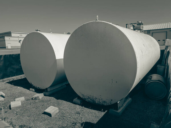 Two large storage tanks sit beside an industrial building under a bright blue sky. The tanks are positioned on concrete bases, surrounded by gravel and a fence.