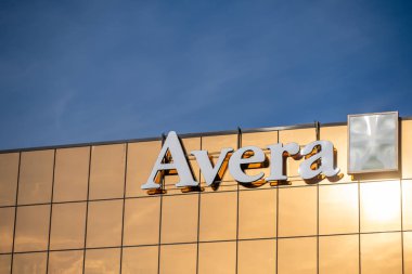 Büyük altın yansıtıcı pencereli parlak bir bina bir Avera işareti sergiliyor. Sahne, öğleden sonra saatlerinde modern mimariyi vurgulayarak açık mavi bir gökyüzü yakalıyor..