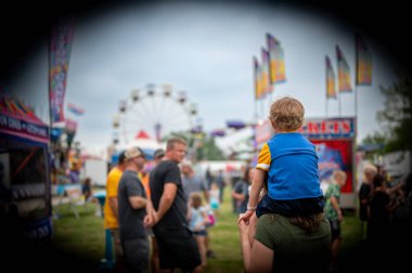 Bir çocuk anne ve babasının omuzlarına tünemiş canlı bir karnavalın manzarasını seyrediyor. Yakınlarda renkli bayraklar dalgalanıyor ve uzaklarda bir dönme dolap beliriyor..