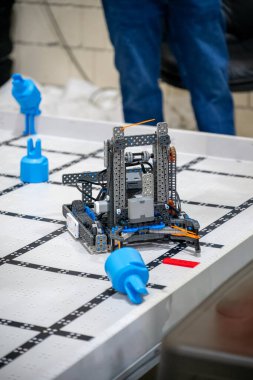 Sınıfta işaretli bir yüzey üzerinde bir robot oturuyor. Öğrenciler robotun masaya konan mavi engelleri aşmaya programlandığını izliyor..