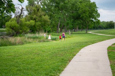 İki yetişkin kaldırım yolunda yürürken, dört çocuk bir parkta bir su kütlesinin yanındadır. Gökyüzü bulutlu ve ağaçlar yeşil. Yaz geldi ve çimenler yemyeşil..