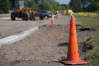 Arka planda ağır makineler ve ekipmanlarla yol yapımı devam ediyor. Güneşli bir günde araçlar geçerken trafik konileri inşaat alanını işaretliyor.