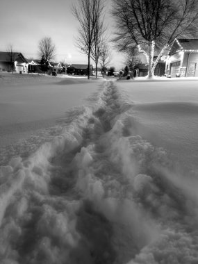 Gece banliyödeki bir mahallede ayak izleri derin karlar arasında bir patikayı işaret ediyor. Evler ve ağaçlar berrak bir gökyüzünün altında serin bir kış manzarası yaratıyor..