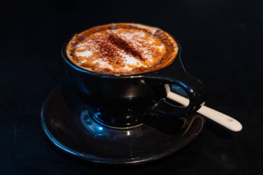 Bir fincan dumanı tüten kahve koyu bir masada oturur. Köpüklü krema ve tarçın serpintisi gibi zengin aromalar havayı doldurur ve lezzetli bir ziyafet yaratır.