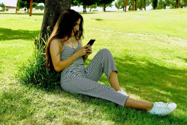 Parkta çimlerin üzerinde oturan akıllı telefonlu bir kız.