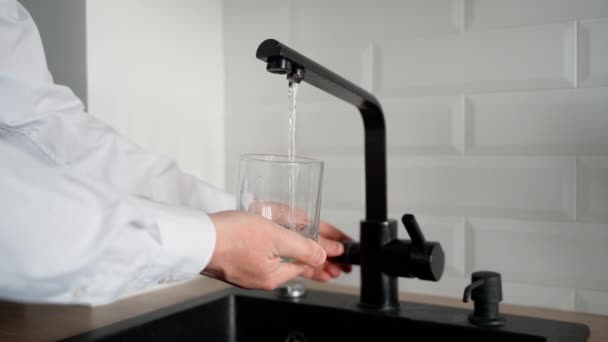 人は台所の飲料水で空のガラスを満たす 水道水は水道水から透明容器にゆっくりと流れます フレームにはシンク タップ 男性用の手とガラスが見えます — ストック動画