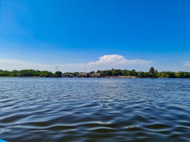 Sakin mavi bir göl, berrak ve parlak bir gökyüzünün altında uzanıyor. Sakin su, açık gökyüzünün yumuşak renklerini yansıtarak büyüleyici bir manzara yaratır. Uzakta, yemyeşil bitki örtüsü ufukta beliriyor, lagünlere doğal güzellik ve huzur katıyor.