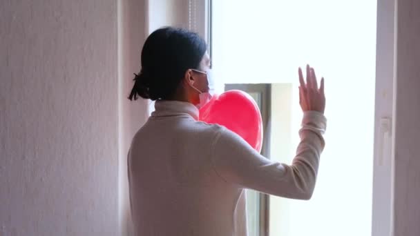 Sad girl in a protective medical mask with a red balloon near the window. — Stock Video