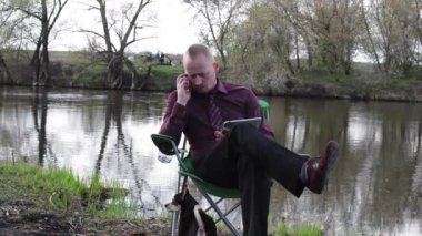 İşadamı oturan ile bir Tablet, telefon konuşmak ve iş için kamp için nehir tarafından zorlanır