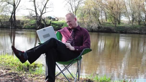 Empresário sentado com uma tabuleta, alegrando-se sorte e é forçado a trabalhar para acampar junto ao rio — Vídeo de Stock