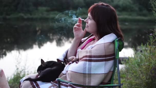 Girl on the Riverbank Drinking Alcohol Out of the Bottle and Smoking a Cigarette. on Lap Dog Lying — Stock Video