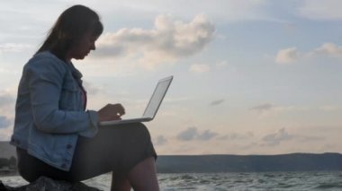 Güneş ışığı ile Laptop ve özlüyor'kayalık kıyısında oturan bir genç bayan. Güçlü dalgalar dayak kayalara karşı