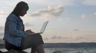 Güneş ışığı ile Laptop ve özlüyor'kayalık kıyısında oturan bir genç bayan. Güçlü dalgalar dayak kayalara karşı