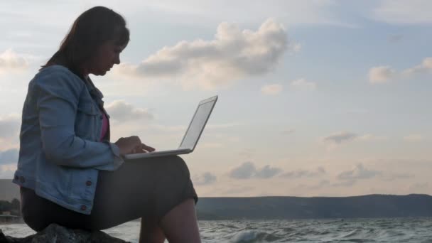 Jonge vrouw zittend op de rotsachtige kust in het zonlicht met Laptop en ongelukken. Sterke golven slaan tegen de rotsen — Stockvideo