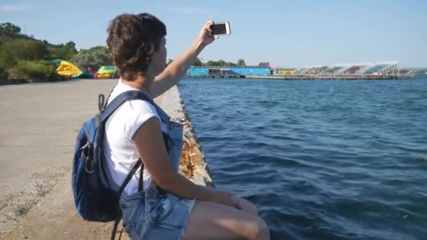 Chica haciendo selfie sentado en un muelle cerca del agua — Vídeos de Stock