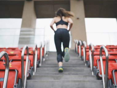 Genç bir kadın spor için stadyuma koşuyor, esniyor, su içiyor.