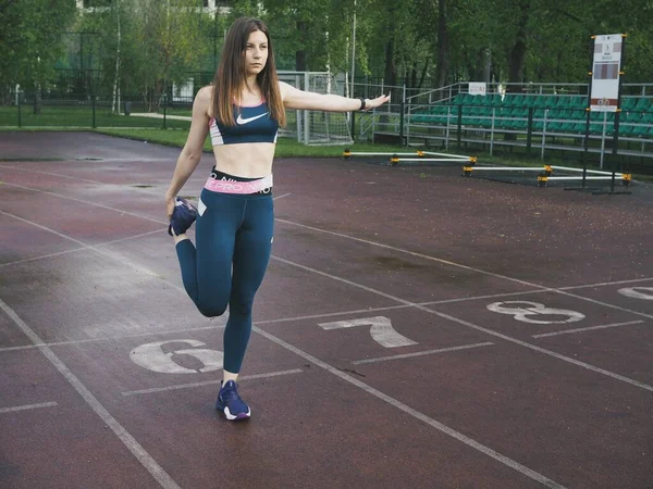 Genç bir kadın spor için stadyuma koşuyor, esniyor, su içiyor.