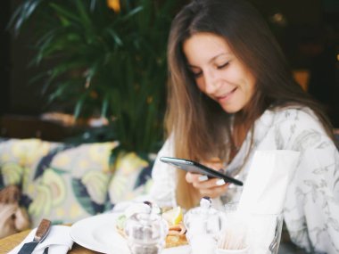 Genç bayan bir kafede telefon kullanıyor. Yemek fotoğrafları çekip telefonda konuşuyorlar.