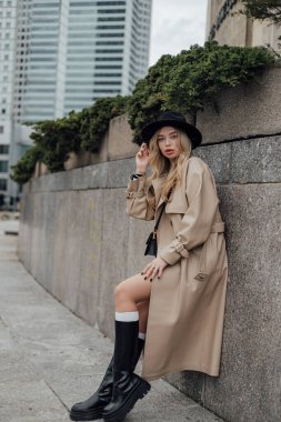 Trençkotlu ve şapkalı genç beyaz kadın şehir ortamında poz veriyor..