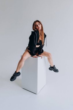 Siyah kapüşonlu, beyaz küpte oturan kendine güvenen bir kadın..