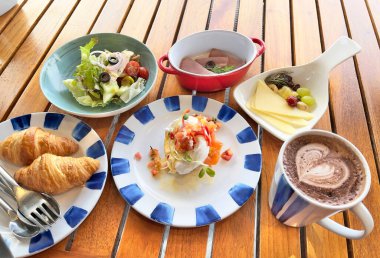 Batı kahvaltı seti: Yumurtalı Benedict, Cesar salatası, Soğuk Pirzola, Peynir tabağı, kruvasan ve ahşap masada sıcak çikolata