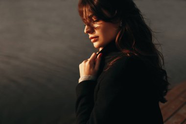 Sıcak giysili düşünceli genç kadın suyun yanında duruyor.