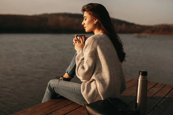 Mujer tranquila con taza de bebida caliente sentado cerca del lago — Foto de Stock