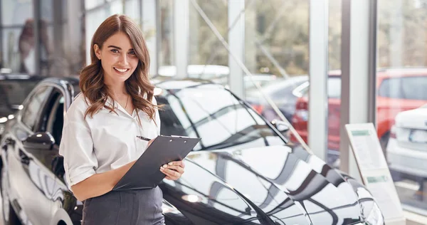 Vendedora positiva com prancheta trabalhando na concessionária de carros — Fotografia de Stock