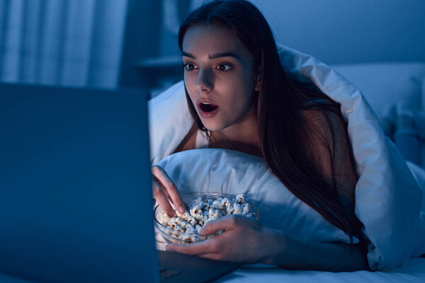 Astonished woman with popcorn watching movie on bed