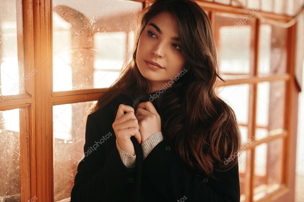 Dreamy woman in warm coat standing near window