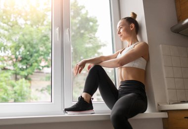 Evde pencere kenarında oturan sporcu kadın.