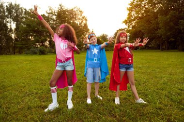 Cheerful diverse children in superhero costumes on summer meadow clipart