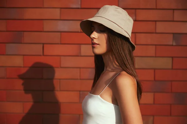 Elegante mujer milenaria en sombrero de pie cerca de la pared de ladrillo —  Fotos de Stock