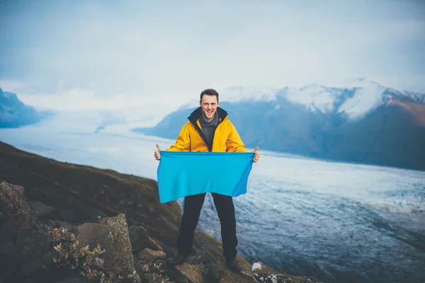 Leende ung man innehar en tom flagga på toppen av berget. — Stockfoto