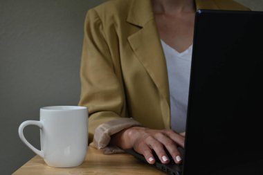Ceketli genç Latin bir kadının yakın plan fotoğrafı. Yanında laptopuyla çalışıyor. Tahta masa üzerinde, fotokopi masası olan bir fincan kahve.