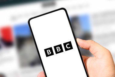 El ele tutuşan akıllı telefon, bulanık haber sayfasında BBC logosu illüstrasyon makalesini gösteriyor.