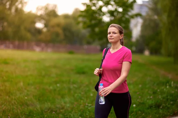 穿着运动服的年轻女子拿着一瓶水在公园里走着 图库图片