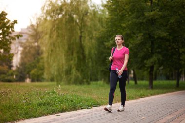 Spor kıyafetli genç bir kadın elinde bir şişe suyla parkta yürüyor.