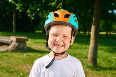Beş yaşında, koruyucu kasklı, kızıl saçlı küçük bir çocuk parkta dikiliyor ve gülümsüyor.